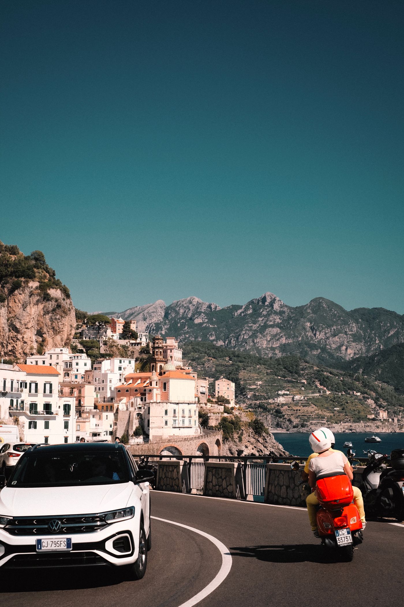 Car on a coastal road by the Mediterranean, a village clinging to steep cliffs in the background
