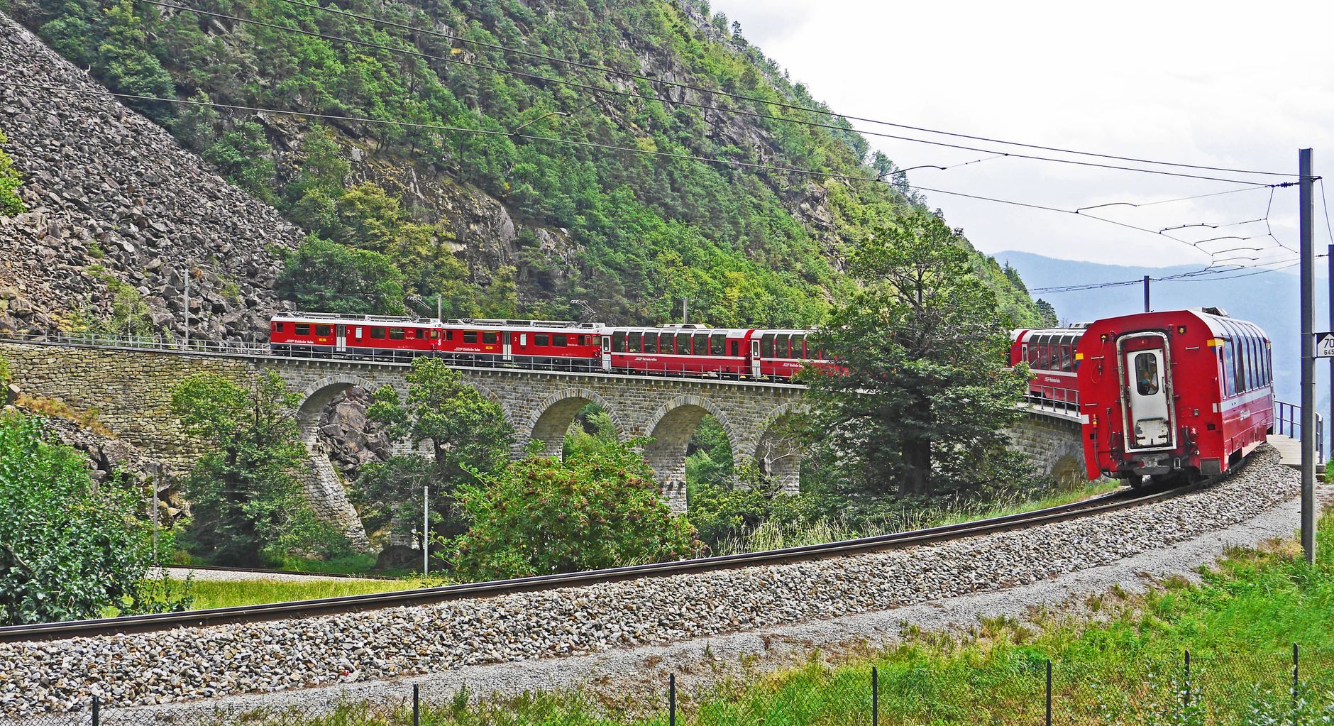 Red narrow-gauge train on a stone viaduct along a forested mountainside