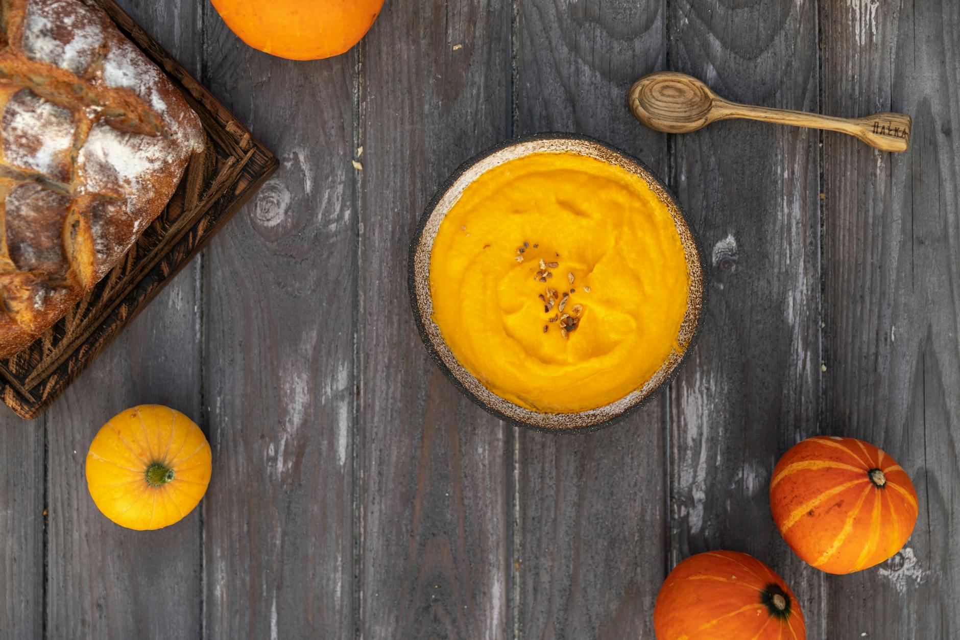 Top view of creamy pumpkin soup, artisan bread, and pumpkins on rustic wooden surface, ideal for autumn food themes.