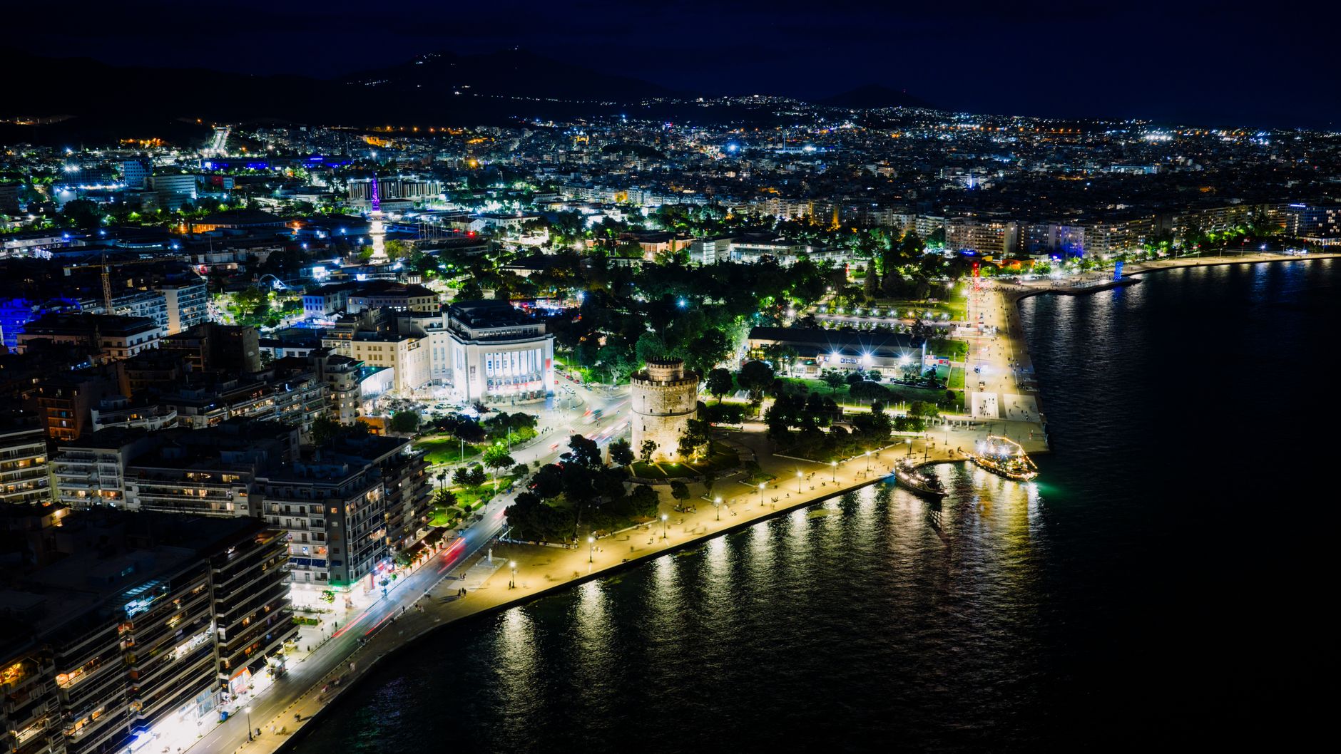 Aerial view of Thessaloniki, Greece at night featuring the illuminated White Tower and waterfront.