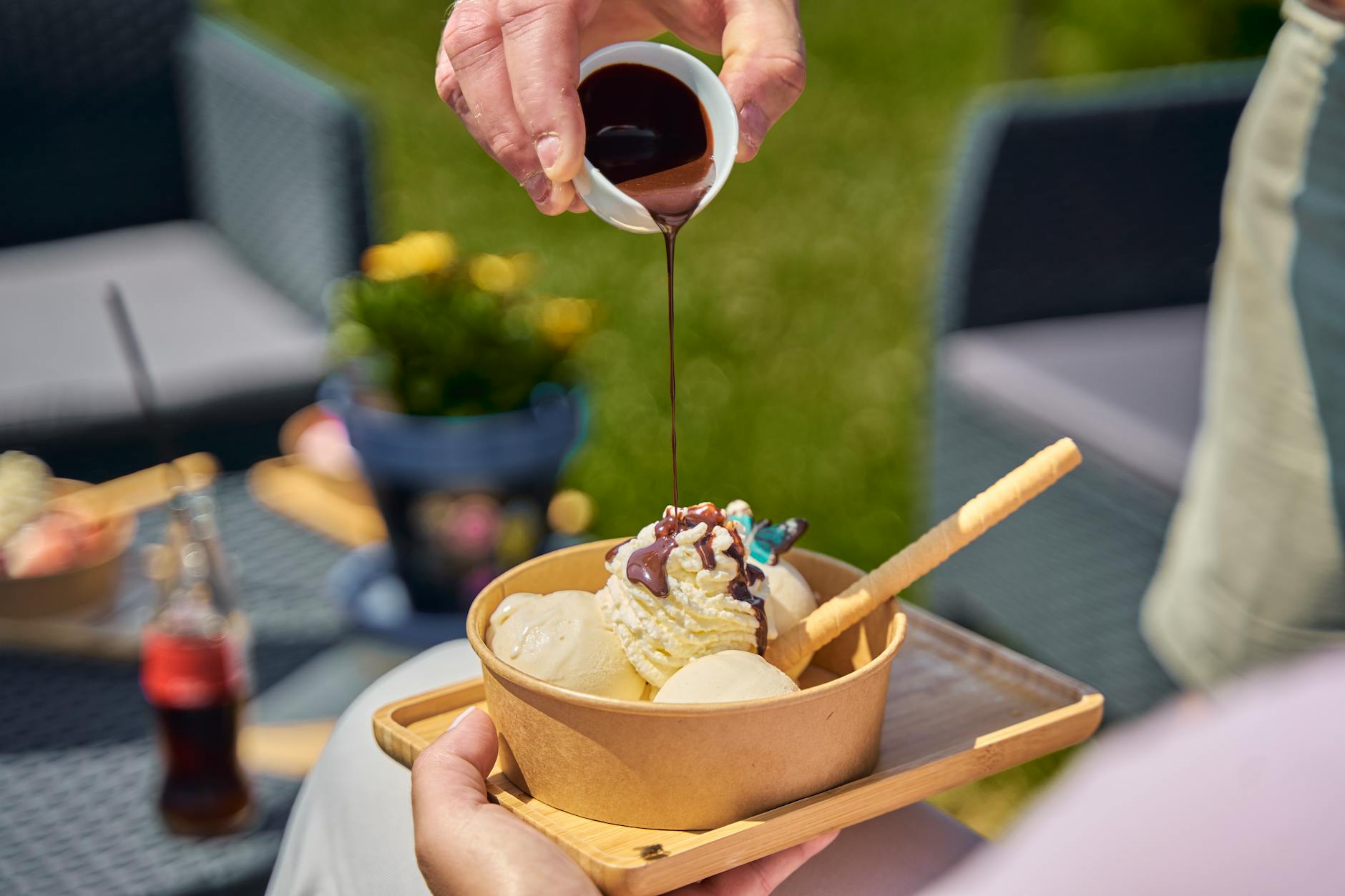 Close-up of chocolate sauce being poured over a bowl of vanilla ice cream outdoors.