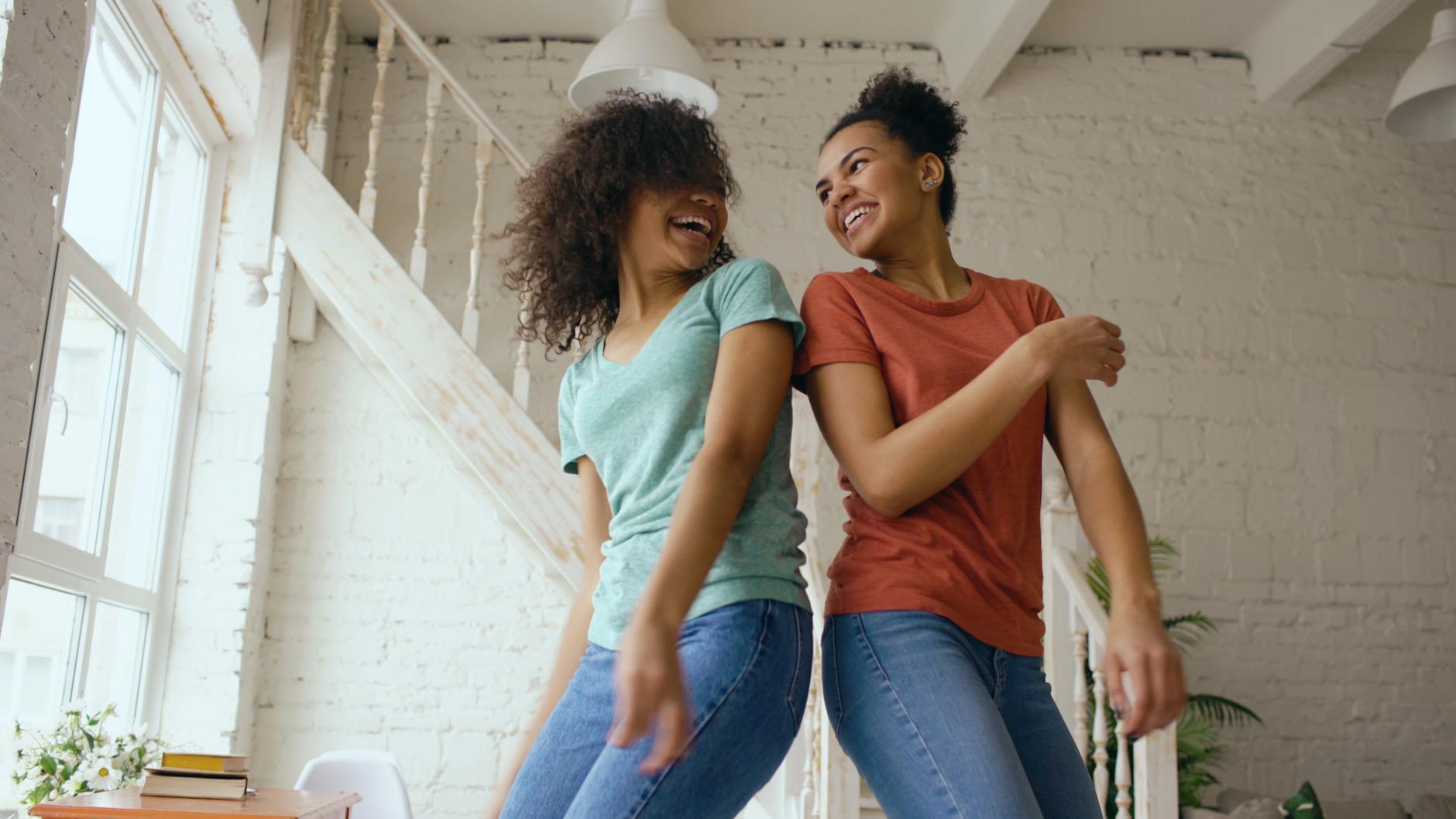 Two young women dancing and laughing indoors by the window, enjoying a joyful moment.