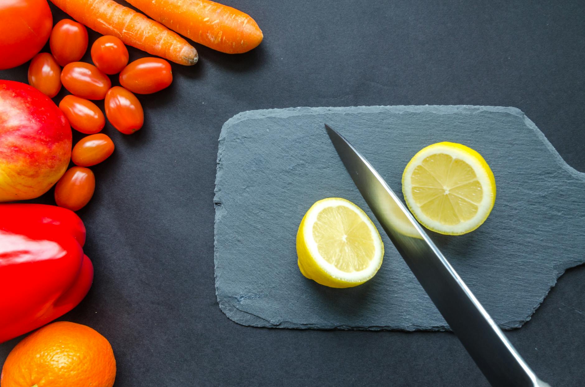 Top view of fresh produce including lemon, carrots, tomato, and peppers on a slate board.