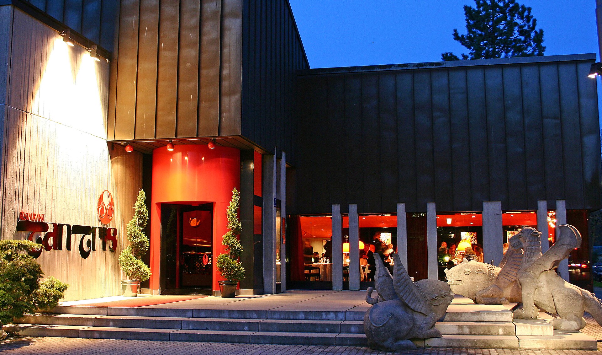 Das Restaurant Tantris in München bei Abenddämmerung: der denkmalgeschützte Bau von Justus Dahinden, warm beleuchtet, mit den Fabeltier-Skulpturen am Eingang