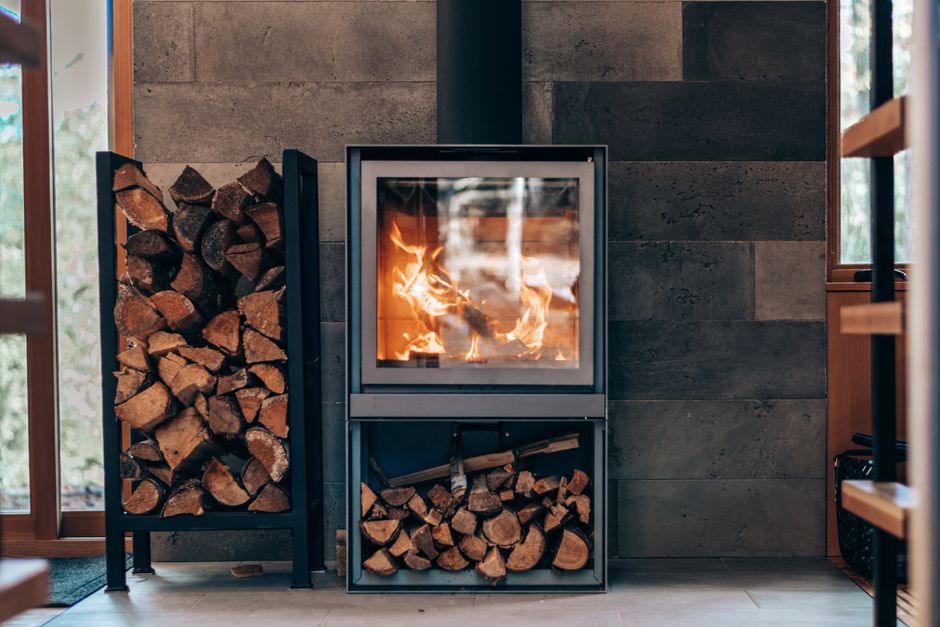You pause by the dark wooden wall, where a wood‑burning stove holds a neat stack of firewood.